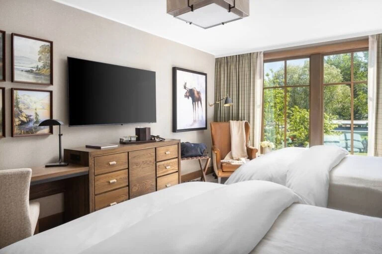 Hotel room with two beds, TV, artwork, and a balcony view of trees.