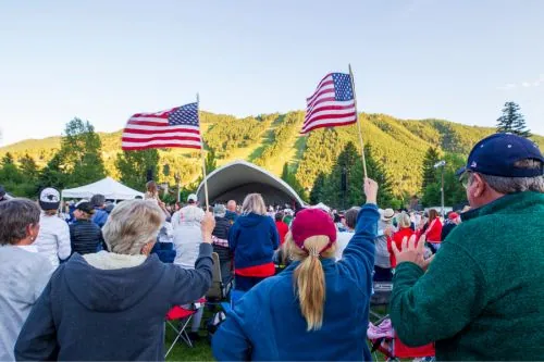 American flags being waved at outdoor event with a crowd of people, stage, and scenic hillside in th.