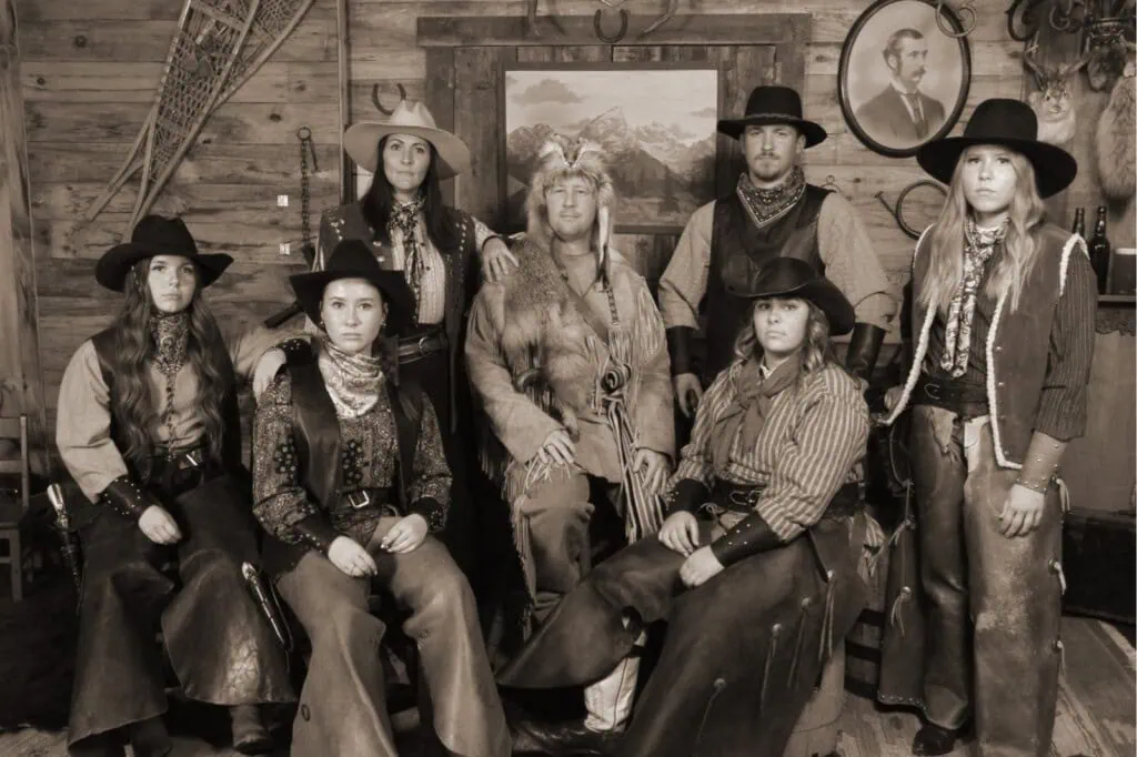 Old West-style group photo of seven people in period costumes, inside a rustic wooden room.