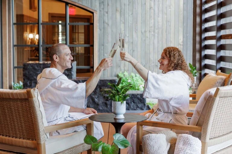 Two women in robes clinking glasses in a cozy spa lounge.