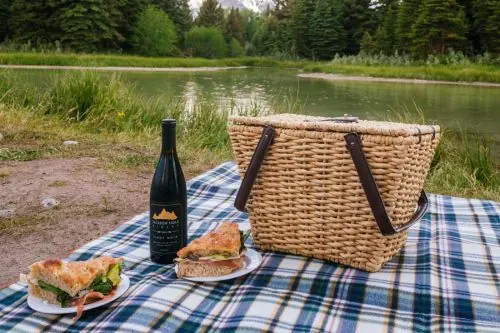 A picnic basket, wine bottle, and sandwiches on a checkered cloth by a lake.