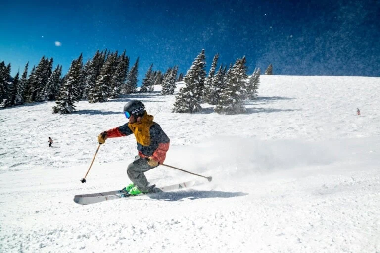 Child skiing downhill on a snowy slope with trees and blue sky in background.