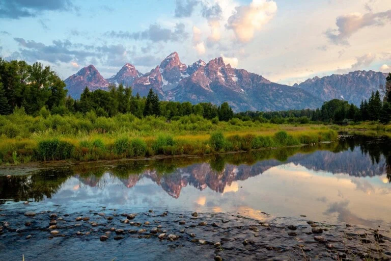 A mountain range reflected in a calm river with greenery in the foreground.