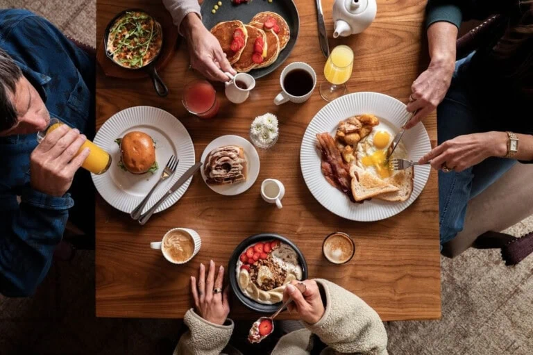 Eggs, bacon, pancakes, toast, and various drinks on a wooden table with people serving and eating.