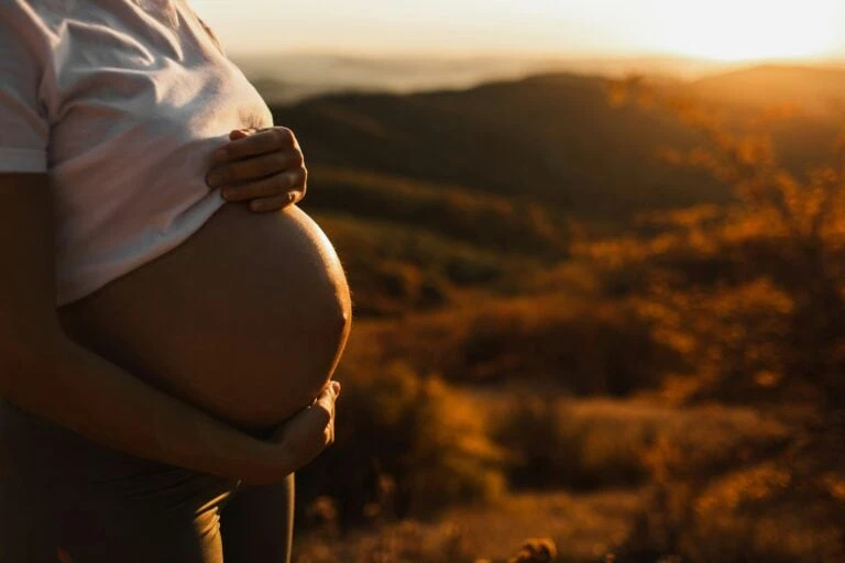 Pregnant woman holding her belly during sunset in a hilly landscape.