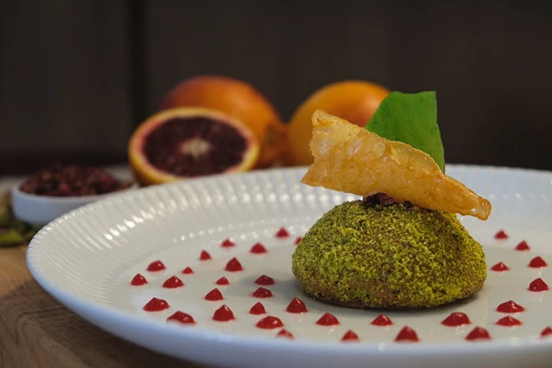 Dessert with green coating, orange slice, and crispy garnish on a white plate.