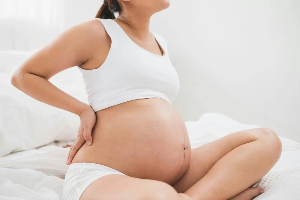 Pregnant woman sitting on bed with hand on her back and belly.