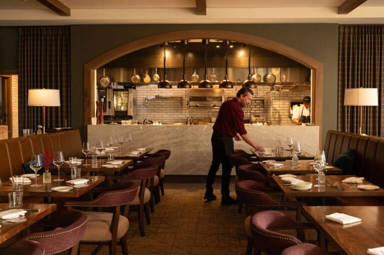 Empty restaurant dining area with a staff member setting tables, open kitchen in background.