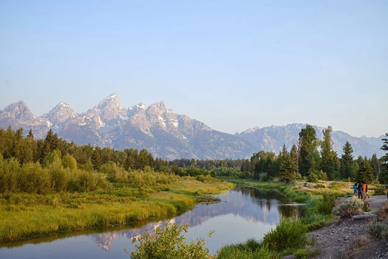 Snow-capped mountains behind a green valley with a river and trees.