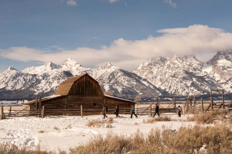 Snow-covered landscape with a wooden barn and mountains in the background.