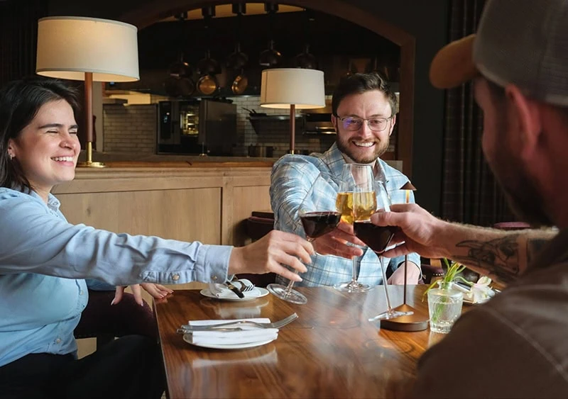 Three people clink glasses of wine and beer at a restaurant table, smiling and enjoying.