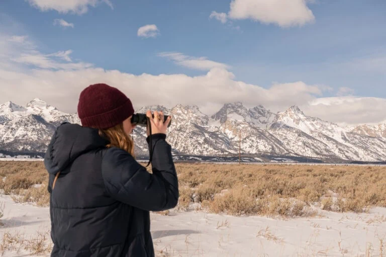 Woman in black jacket and red beanie taking photos of snowy mountains.