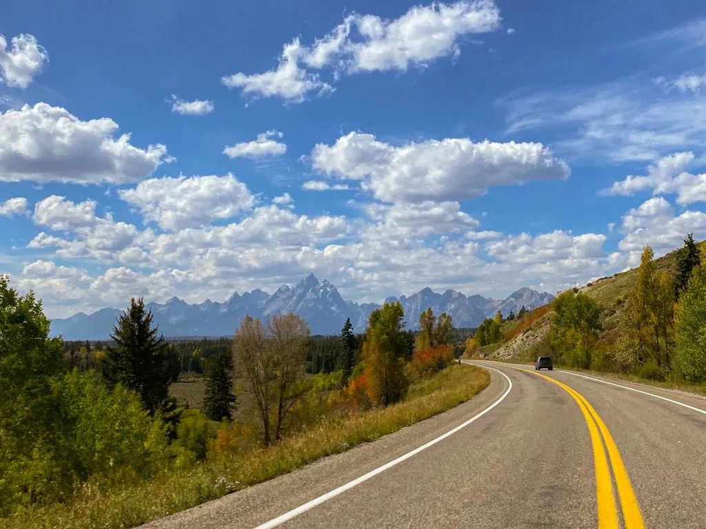 Scenic mountain road with yellow lines, trees, and mountains in the distance under a partly cloudy.