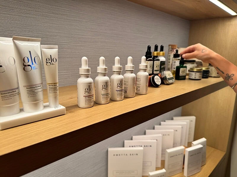 A hand reaching for skincare products on a wooden shelf, including Glo and Ametsa Beki items.