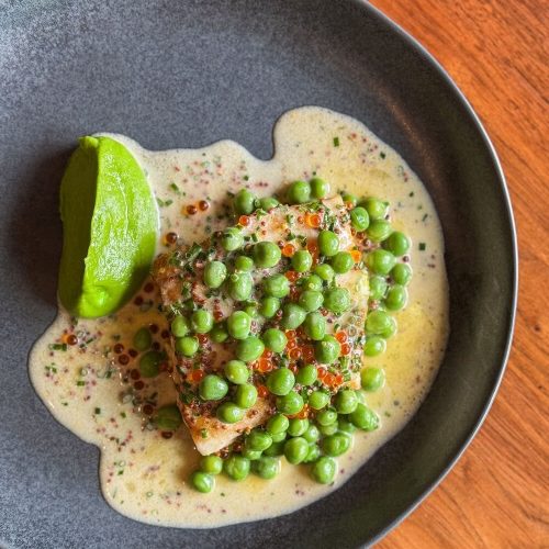 Fish fillet with green peas and lime wedge in creamy sauce on black plate.