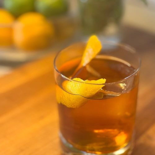 A glass of amber-colored cocktail with lemon peel garnish on a wooden surface.