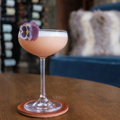 Pink cocktail in a coupe glass with a purple flower garnish.