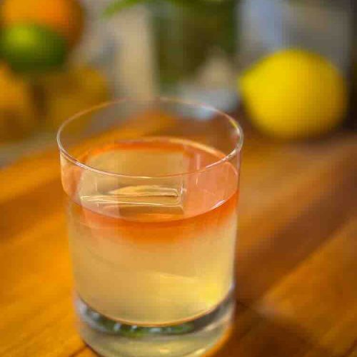 A glass of layered cocktail with a pink top and yellowish bottom on a wooden table.