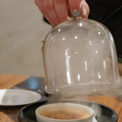 Person lifting a glass cover over a small dish on a black plate.