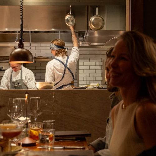 Kitchen staff preparing food in a restaurant with a smiling woman in the foreground.