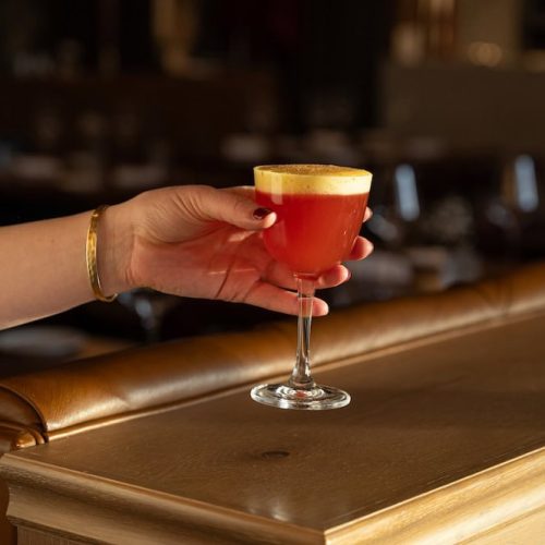 A hand holding a cocktail glass with a layered drink in a bar setting.