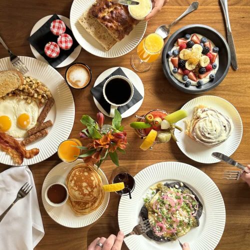 Assorted breakfast foods on a wooden table including pancakes, eggs, fruit, and coffee.