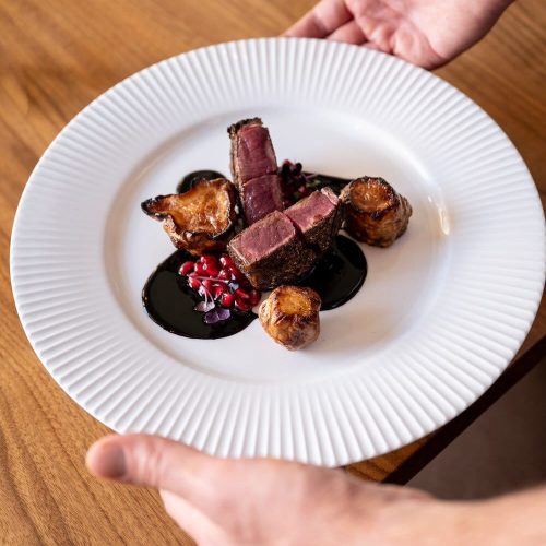 Chef's hand presenting a plated dish with sliced steak, roasted vegetables, and sauce.