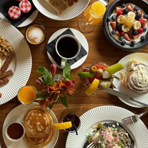 Breakfast table with eggs, bacon, pancakes, fruit, and coffee.