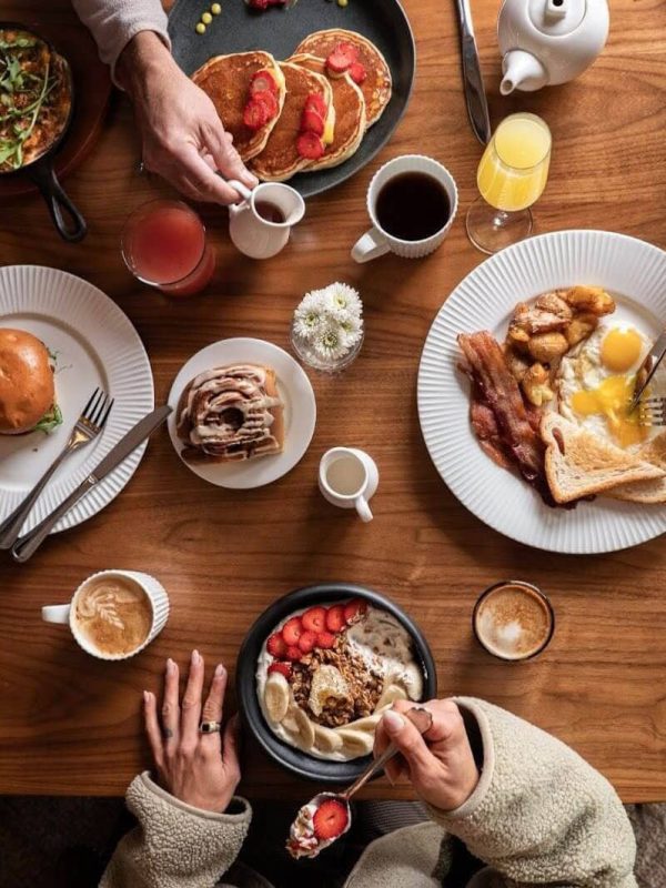 Eggs, bacon, pancakes, toast, and various drinks on a wooden table with people serving and eating.