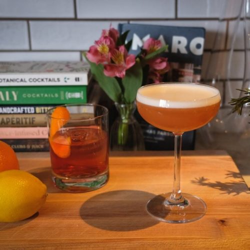 Two cocktails on a wooden bar with books, flowers, and plants in the background.
